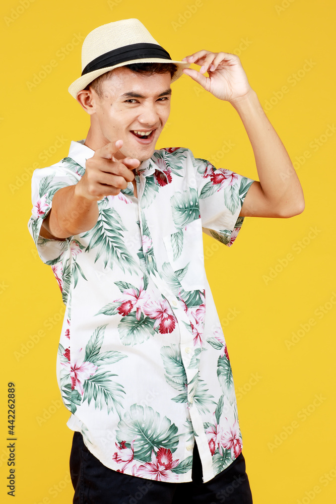 Young attractive Asian man in white Hawaiian shirt grabbing his hat and pointing and smiling against yellow background. Concept for beach vacation holiday
