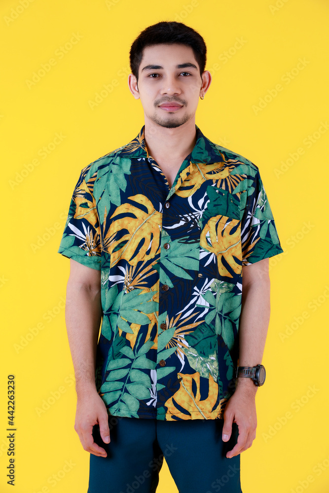 Fototapeta premium Handsome young Asian man in colorful Hawaiian shirt standing looking to camera. Half body studio portrait on yellow background. Relaxed and happy people in summer concept