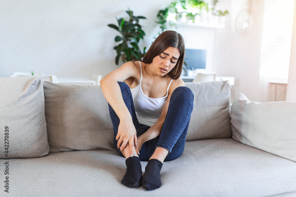 Hands massaging swollen foot while sitting on sofa during the day at home. Photo of Young Caucasian woman suffering from pain in leg. Woman massaging her legs after all day at work in office Hands massaging swollen foot while sitting on sofa during the day at home. Photo of Young Caucasian woman suffering from pain in leg. Woman massaging her legs after all day at work in office