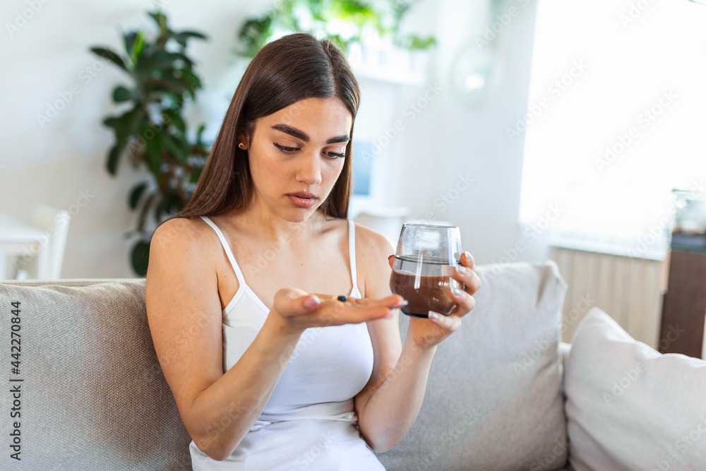 Concept of suffering from terrible pain. Beautiful, sad, upset, unhappy, troubled, weak woman with black hair, wearing casual clothes is sitting on a sofa and taking a pill Concept of suffering from terrible pain. Beautiful, sad, upset, unhappy, troubled, weak woman with black hair, wearing casual clothes is sitting on a sofa and taking a pill