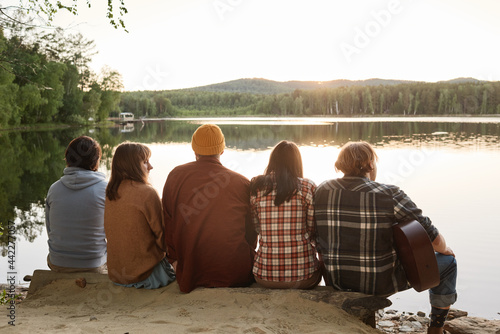 Rear view of friends playing guitar while sitting on the edge of the bank and enjoying the view of beautiful lake outdoors