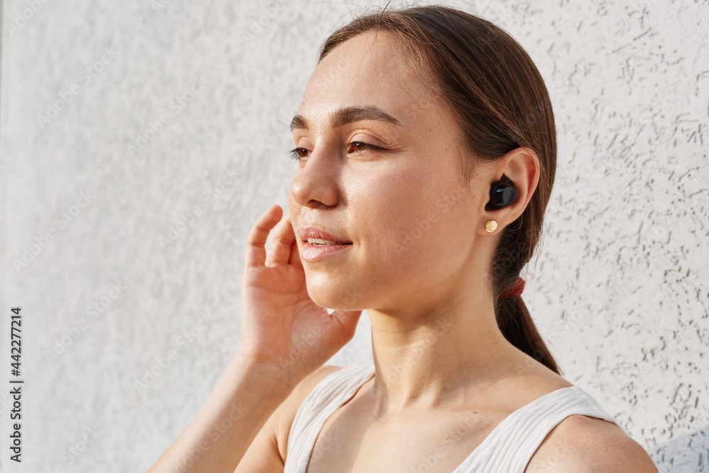 Side view portrait of good looking female with dark hair looking away and touching airpods, wearing white top, standing isolated over gray background outdoor. Side view portrait of good looking female with dark hair looking away and touching airpods, wearing white top, standing isolated over gray background outdoor.