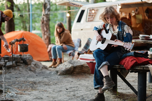 Young woman playing guitar while resting on a camp with her friends in the forest