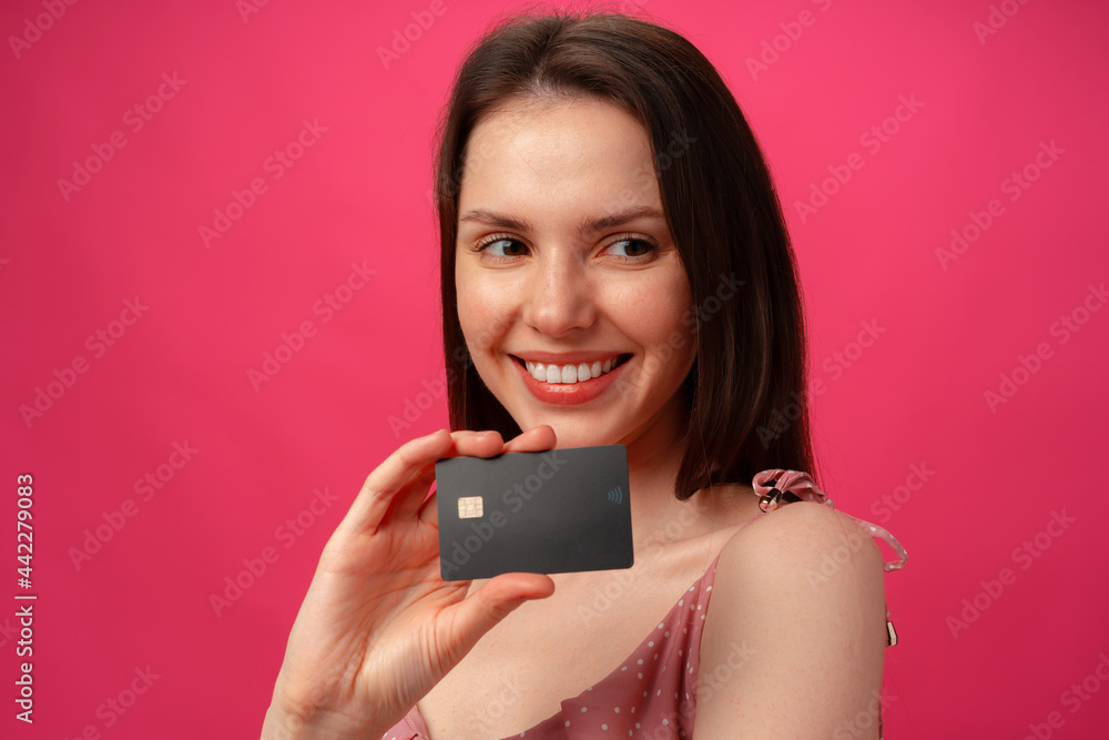 Attractive smiling young woman holding black credit card against pink studio background Attractive smiling young woman holding black credit card against pink studio background