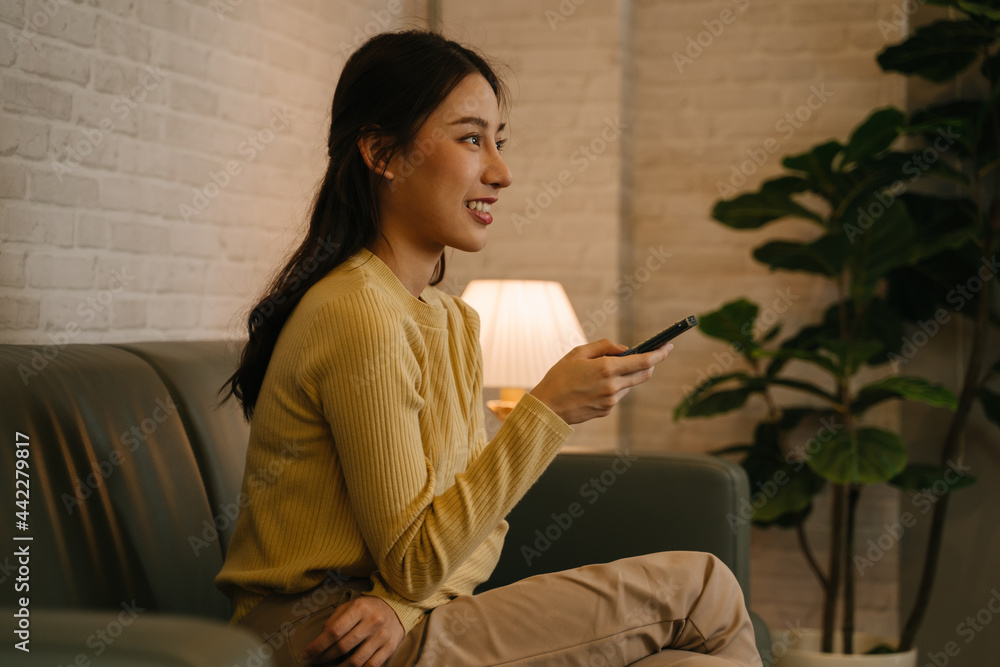 Side view of young happy Asian woman holding tv remote control changing channel while sitting on couch at home during late night Side view of young happy Asian woman holding tv remote control changing channel while sitting on couch at home during late night
