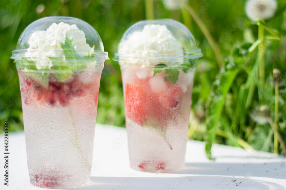 Cocktail with watermelon slices and ice cubes in a plastic cup, with a white flower. On a white wooden background, in the grass. Summer tropical refreshing drink. Cocktail with watermelon slices and ice cubes in a plastic cup, with a white flower. On a white wooden background, in the grass. Summer tropical refreshing drink.