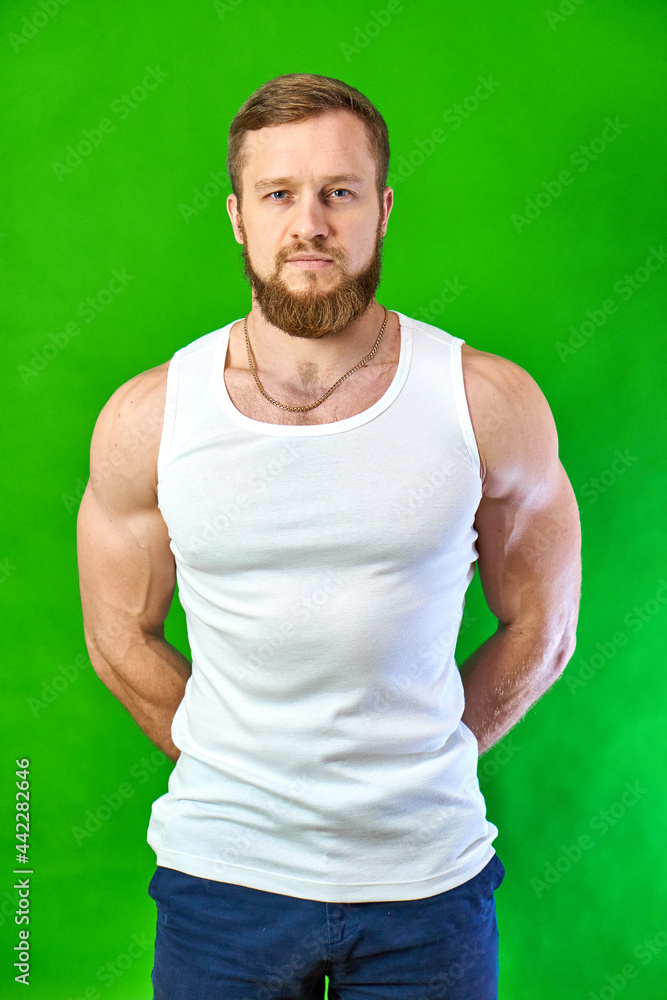 Fototapeta premium The red-haired man looks at the footage. Serious sportsman face with muscles. A Caucasian man with a beard stands on a green background in a white T-shirt and jeans.