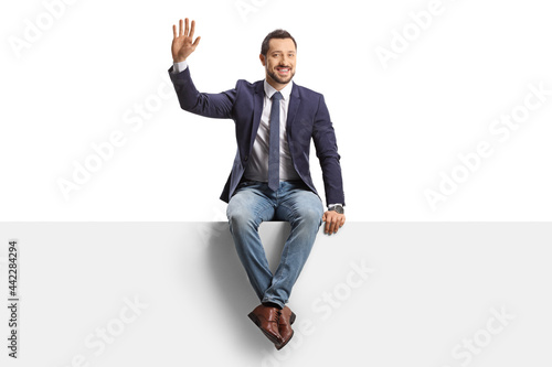 Full length portrait of a man in jeans and tie sitting on a blank panel and waving