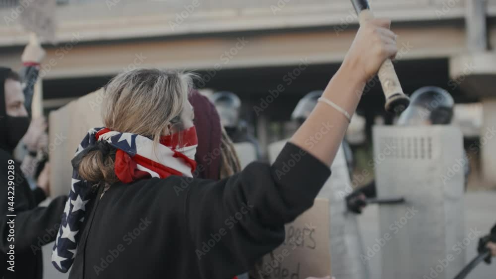 Slowmo shot of masked young people with signs and baseball bat protesting outside while riot police with shields blocking street Slowmo shot of masked young people with signs and baseball bat protesting outside while riot police with shields blocking street