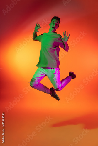 Portrait of Caucasian young man jumping, dancing isolated on orange studio background in neon light. Concept of human emotions, facial expression.