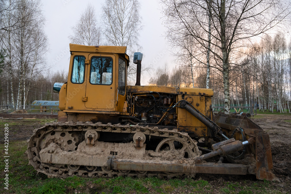 An old rusty yellow tractor among the trees in the open air on a driverless farm. Agricultural machinery An old rusty yellow tractor among the trees in the open air on a driverless farm. Agricultural machinery