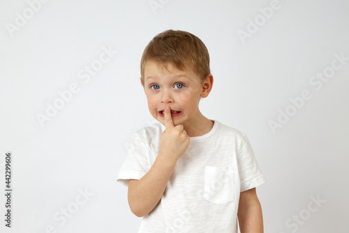 Cute little boy in casual clothing standing on white background of studio and putting index finger on lips while looking at camera. The child keeps a secret