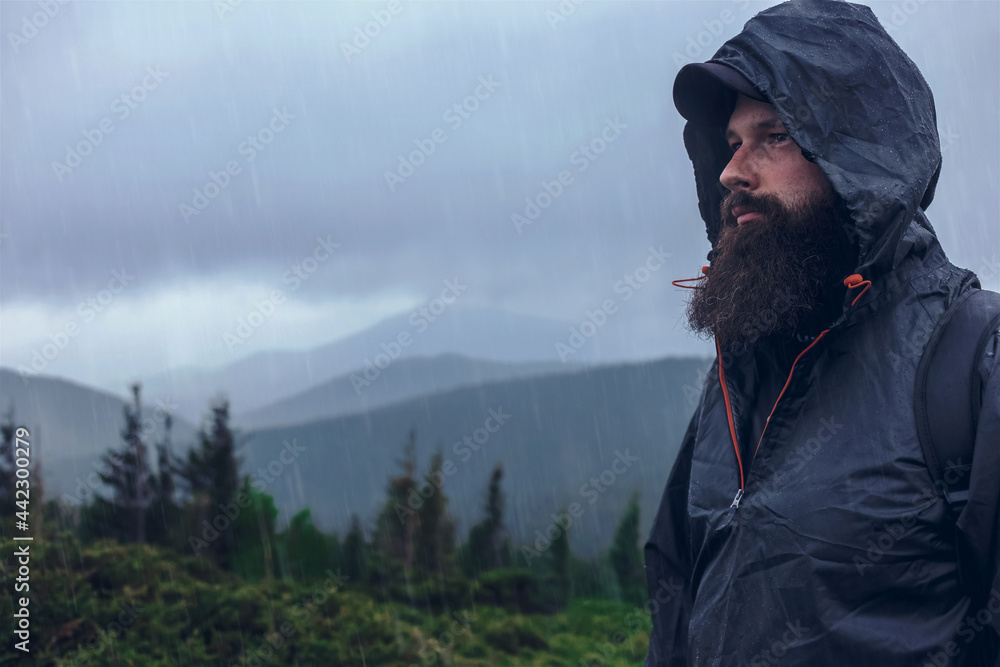 Handsome serious hiker bearded man on a mountain hill in a waterproof jacket under the summer rain. Mountain top against cloudy sky on natural background, copy space Handsome serious hiker bearded man on a mountain hill in a waterproof jacket under the summer rain. Mountain top against cloudy sky on natural background, copy space