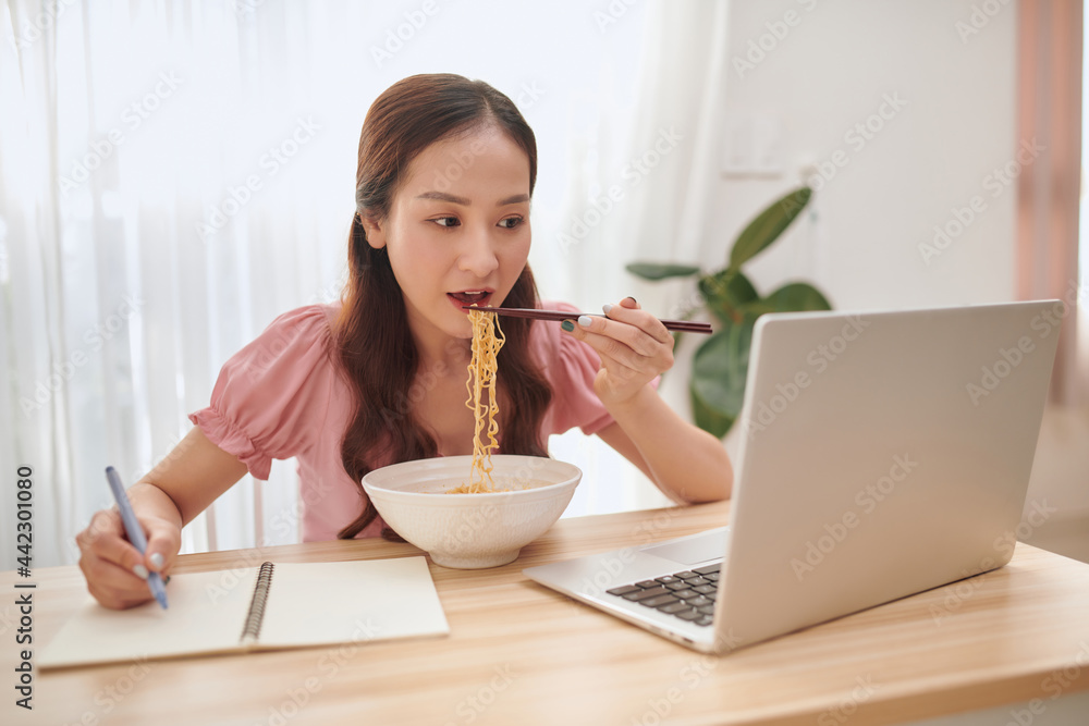 Young Asian woman eating noodles and working with laptop at home. Young Asian woman eating noodles and working with laptop at home.