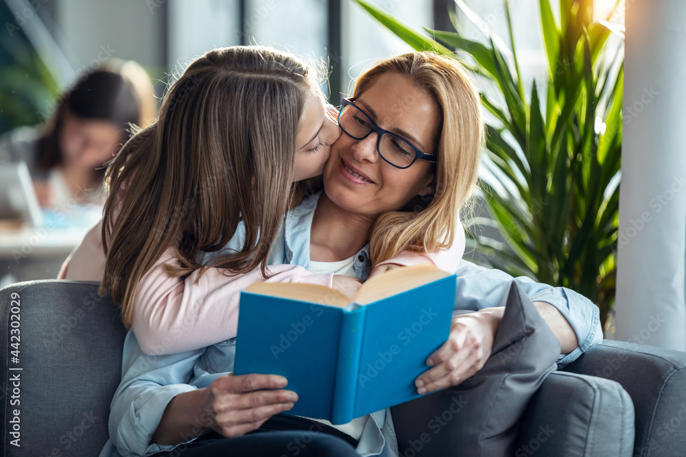 © nenetus - Mature beautiful woman reading a book while her daughter hugging in living room at home.