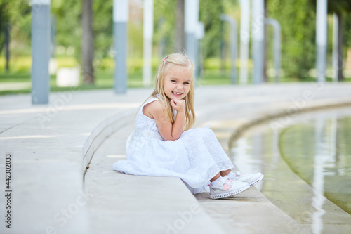 A little blonde girl in a white dress is sitting by a pond in the Park.