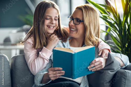 Mature beautiful woman reading a book while her daughter hugging in living room at home.