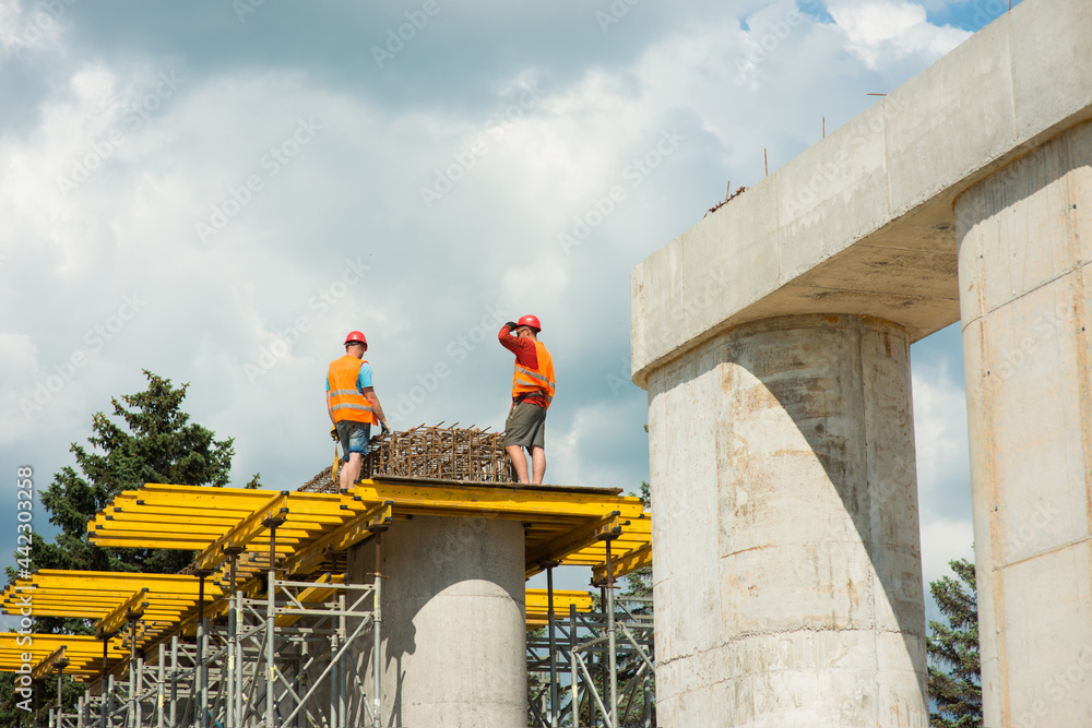 Construction workers wearing safety helmets install reinforced frames on concrete columns while building a road bridge.
