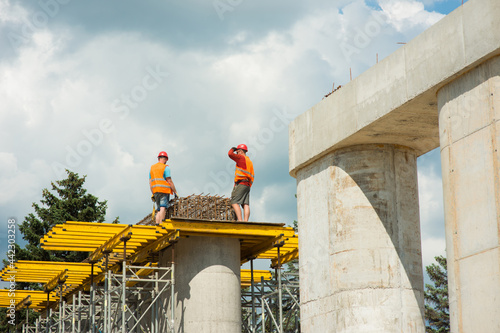 Obraz na płótnie Construction workers wearing safety helmets install reinforced frames on concrete columns while building a road bridge. w salonie