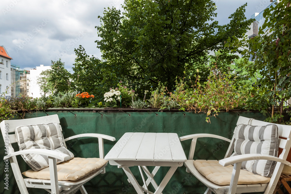 White wooden furniture garden set decorates a green balcony of a city apartment in Germany. Terrace with greenery, wooden table and chairs for two