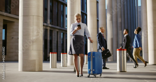 Young businesswoman with suitcase using smartphone waiting for taxi outside airport building