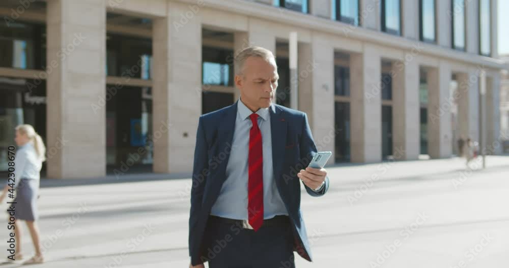 Middle-aged entrepreneur walking outside office building using smartphone Middle-aged entrepreneur walking outside office building using smartphone