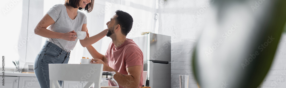 Obraz premium young couple gently touching each other and holding cups with coffee in kitchen, banner