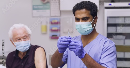 A Mid Shot Of An Indian Male Medical Nurse Administering A Covid-19, SARS-CoV-2 Vaccine Injection With A Needle and Syringe, To An Elderly Man. Wearing Safety Gloves And Protective Mask