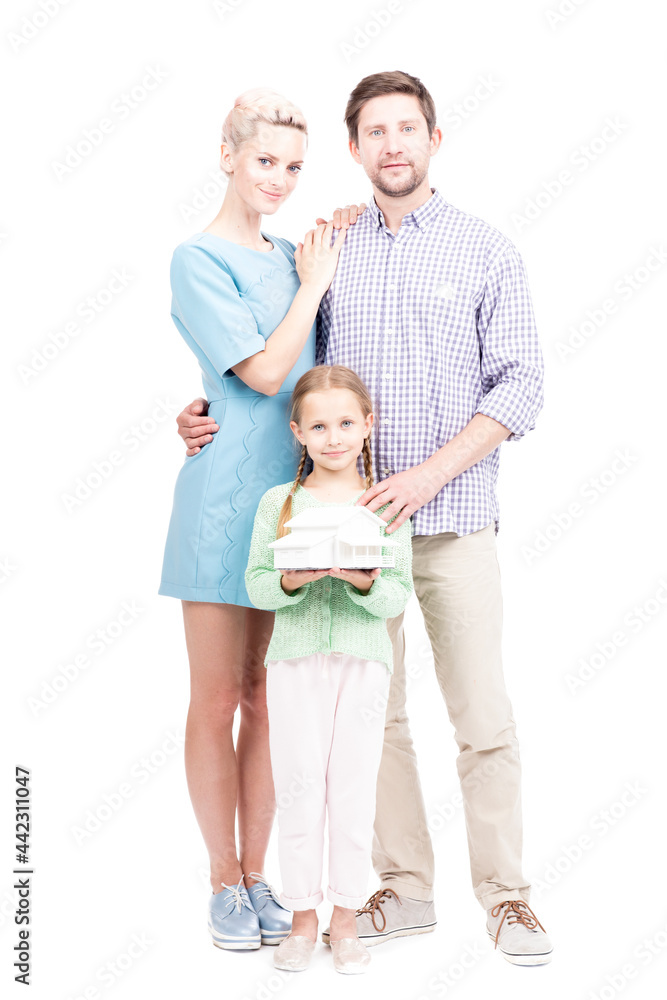 Vertical full length shot of modern family with little daughter standing together, girl holding house model looking at camera, white background