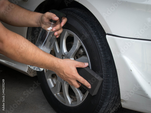 A man's hand holding a spray bottle of wheel cleaner and the other hand is holding a black sponge. wiping the edge of the tire © Itthiphon