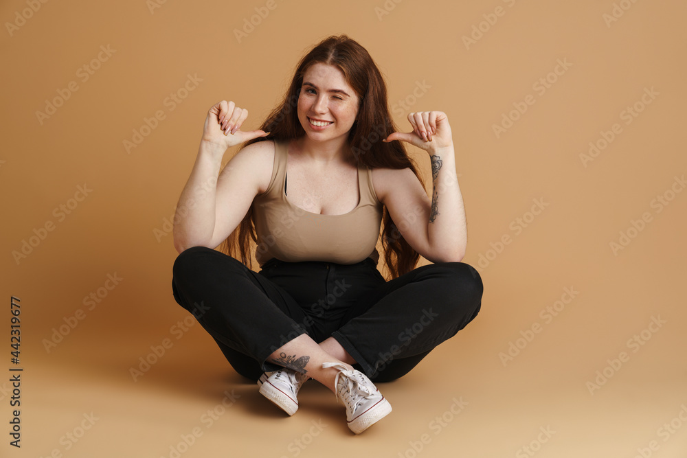 Young ginger woman pointing finger at herself while sitting on floor