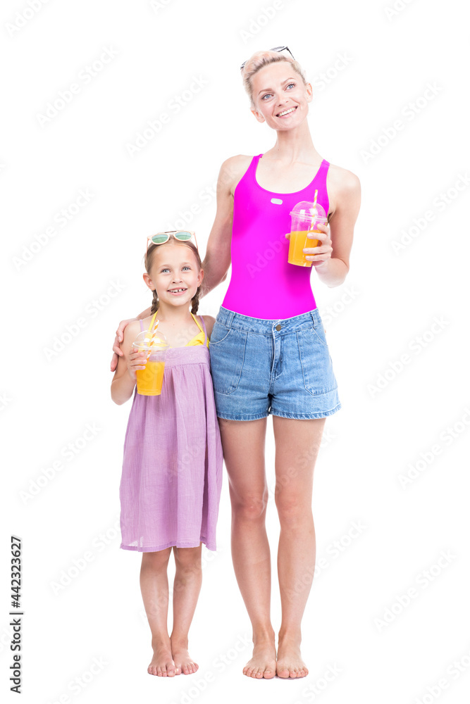 Vertical full length studio portrait of modern Caucasian woman and girl wearing summer clothes standing together drinking fresh orange juice Vertical full length studio portrait of modern Caucasian woman and girl wearing summer clothes standing together drinking fresh orange juice