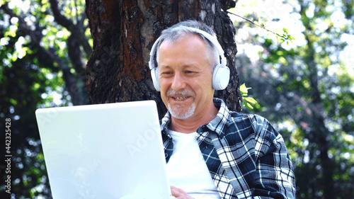 An elderly gray-haired man with a beard in white headphones enjoys music and nature, sitting with a laptop in an open city park on a sunny summer day.