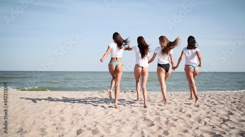 Group of happy female friends undress running into sea water, throw shirts on the beach. Cheerful girls in bikini having fun on shore near ocean. Young women go to swim and bathe. 120 fps slow motion.