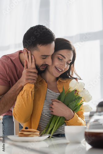 young man hugging girlfriend with bouquet of flowers and closed eyes in kitchen