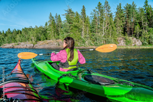 Kayaking women holding paddles horizontally while passing close another kayaking person. She wears magenta jacket and green safety vest. Summer pine tree forest and stones. Umea river, Sweden © Alexandre Patchine