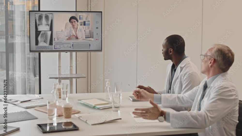 Medium slowmo of two professional multiethnic radiologists sitting at table in conference room having video conversation with female colleague discussing chest x-ray of patient Medium slowmo of two professional multiethnic radiologists sitting at table in conference room having video conversation with female colleague discussing chest x-ray of patient