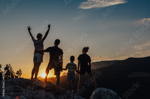 Silhouettes of happy family of four relaxing together outdoors in nature at night. Serene view of dreaming people holding hands and looking at sun