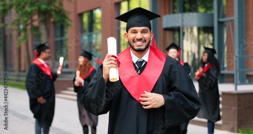 School graduate man in academic gown and hat looking at the camera with happy smile. Happy multiracial man rejoicing near his university or school. Graduation concept