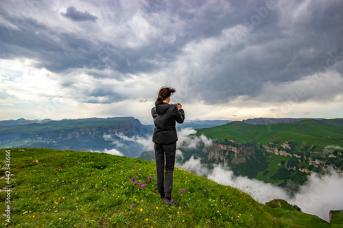 A girl stands on the edge of a cliff and takes pictures of a beautiful view. View from the back. Rest in the mountains. The concept of active recreation. Tourism in the highlands. Rest in solitude.