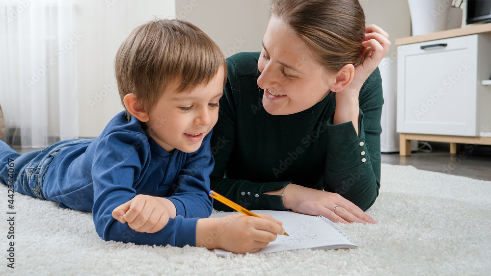 Smiling boy lying on floor and doing homework with his mother. Concept of child education, development and happy parenting. Smiling boy lying on floor and doing homework with his mother. Concept of child education, development and happy parenting.