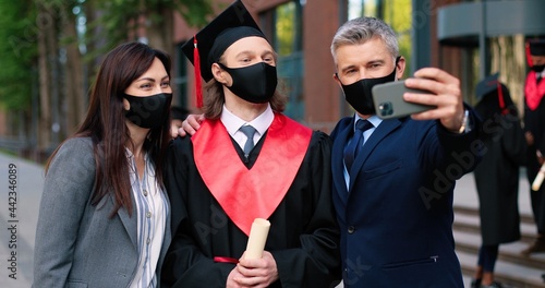 Family portrait. Waist up portrait view of the happy parents wearing protective masks making selfie with their son holding diploma and looking to the camera