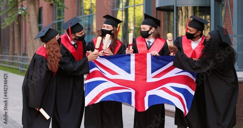 Wallpaper Mural Young graduated people holding their graduation degree convocation ceremony and embracing while posing towards the camera and holding flag of Great Britain with background of academy Torontodigital.ca