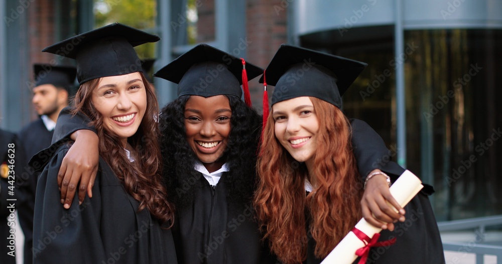 Obraz premium Happy group of mature students on graduation day embracing with each other. Three best girl friends in academic gowns and caps hugging in front of the camera
