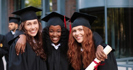 Happy group of mature students on graduation day embracing with each other. Three best girl friends in academic gowns and caps hugging in front of the camera