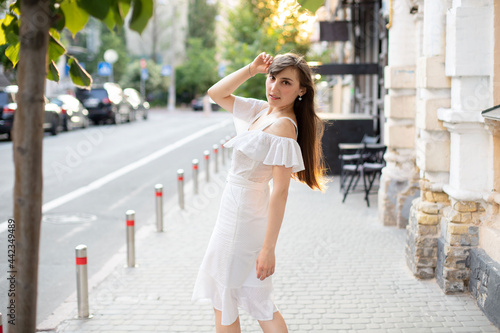 beautiful girl with brown hair in a white dress walks down the street on the background of the road in summer