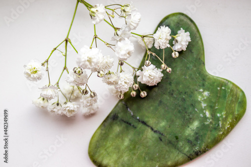A Gypsophila bunch of flowers and a massage face tool, on a white background. Green color gua sha massager. Little white flowers. SPA beauty treatment concept. Facial massager with jade stone, anti-ag