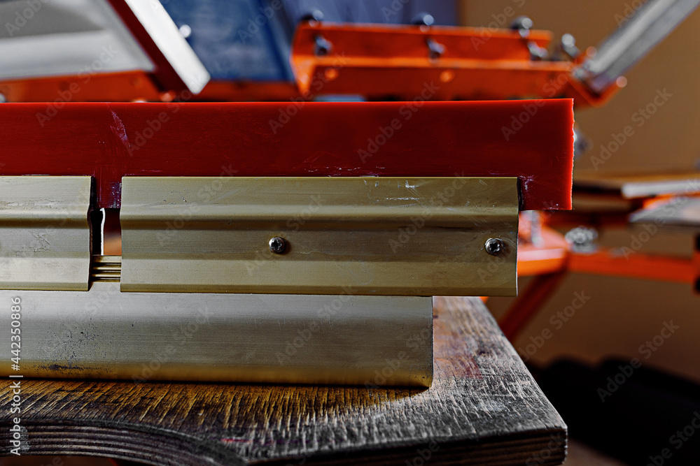 selective focus photo of a squeegee on wooden shelve of the print screening apparatus. serigraphy production. printing images on t-shirts by silkscreen method in a design studio selective focus photo of a squeegee on wooden shelve of the print screening apparatus. serigraphy production. printing images on t-shirts by silkscreen method in a design studio