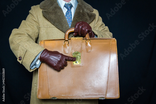 Portrait of Man in Fur Coat and Leather Gloves Pointing to Combination Lock on Briefcase on Black Background. Identity Theft Protection. Vintage Film Noir Secret Agent Spy Hero. photo