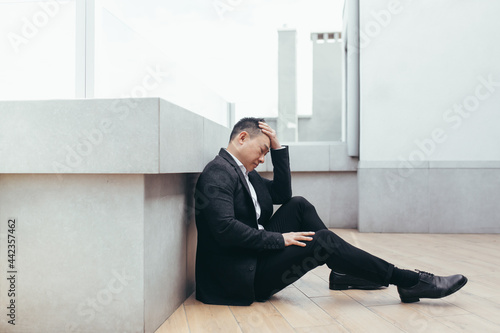 young asian depressed man sitting on outdoors street near office business center building. The upset male businessman lost his job due to the financial crisis and quarantine. Businessman has problems